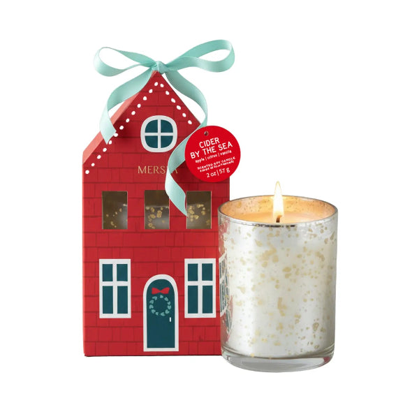 A personalized red house-shaped candle holder with a mint green ribbon bow next to a lit white candle in a gold-speckled glass holder. The house features cutout windows and the name 'MERS' printed on the front, with a red circular tag reading 'CIDER BY THE 364'.