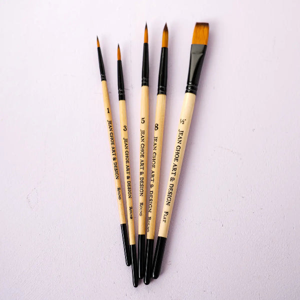 Five paint brushes with light wood handles and black ferrules, arranged in a fan shape on a light pink textured background. The handles are printed with the text 'JEAN CHOE ART & DESIGN'.
