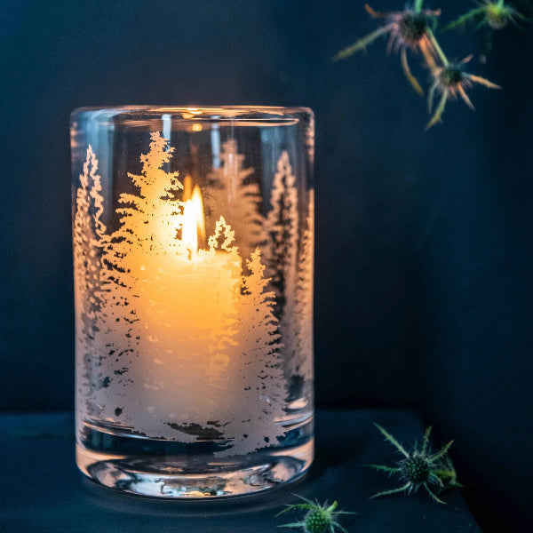 A lit tealight candle inside a clear glass holder etched with a winter forest pattern of pine trees, set against a dark blue background with decorative thistle flowers.