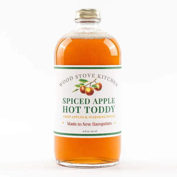 A clear glass bottle of Wood Stove Kitchen Spiced Apple Hot Toddy filled with amber-orange liquid, featuring a white label with green and red text and graphics, and a gold screw cap, on a white background.