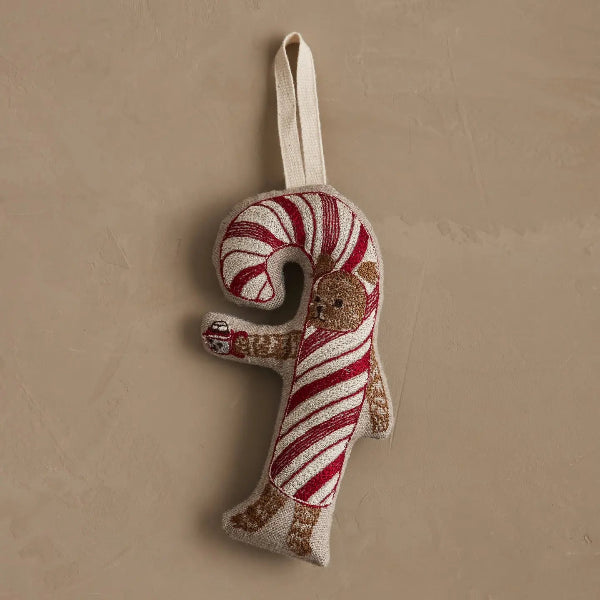 A handmade Christmas ornament shaped like a candy cane, featuring red and white striped fabric with a small embroidered dog and a beige hanging loop, set against a neutral beige background.