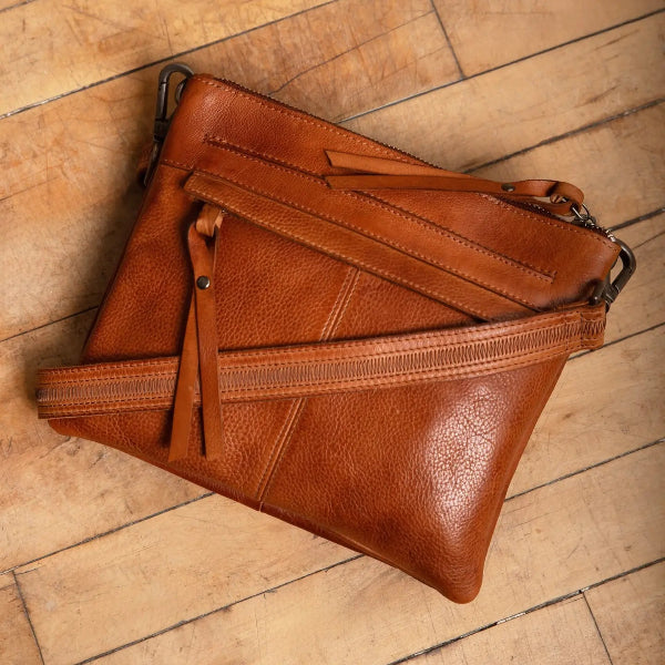 A brown leather clutch bag with a zipper closure and a leather strap detail, displayed diagonally on a light-colored wooden plank floor.