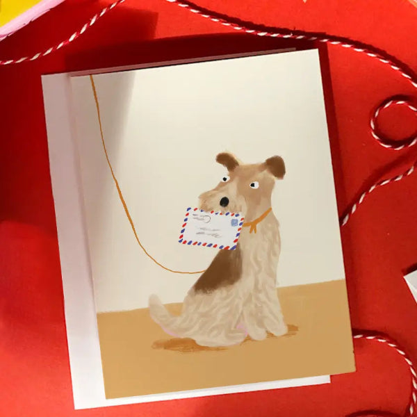 A greeting card featuring an illustration of a wirehaired dog holding a postcard, displayed on a red textured fabric background with white cord.