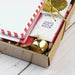 Gift box with 'North Pole Snow' card, gold bell, and red and white striped envelope on a wooden surface.