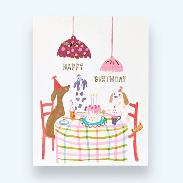 A square birthday greeting card featuring a whimsical illustration of two dogs sitting at a table with a cake, chairs, hanging lamps, and the words “HAPPY BIRTHDAY” in gold lettering, shown against a light blue background.