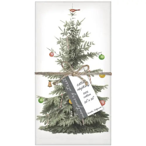 A rectangular white napkin featuring a vintage-style illustration of a Christmas tree with green needles, colorful ornaments, a small bird topper, and a wrapped tag reading 'Cotton napkins 100% cotton 20 x 20"'.