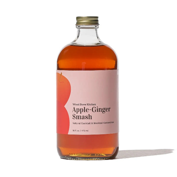 A bottle of Wood Shore Kitchens Apple-Ginger Smash cocktail and mocktail concentrate, featuring a light pink label with a red apple graphic and a gold metal cap, set against a plain white background.