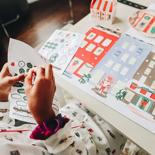 A person's hands are shown assembling a Christmas-themed paper craft or decoration on a white table, with colorful printed paper pieces featuring Santa, trees, and holiday designs.