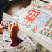 A person's hands are shown assembling a Christmas-themed paper craft or decoration on a white table, with colorful printed paper pieces featuring Santa, trees, and holiday designs.
