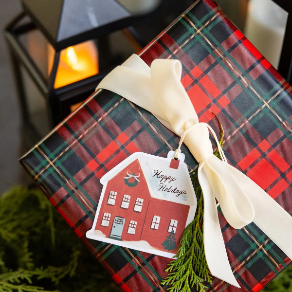 A gift box wrapped in red, green, and black plaid paper with a cream ribbon bow and a holiday tag that says 'Happy Holidays' in front of a softly blurred lantern and greenery.