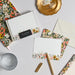 A styled flat-lay of floral stationery items on a light gray surface, including a boxed notepad set, envelopes, a gold pen, a small gold clip, and a glass dish. The stationery features multicolor floral patterns with green, pink, orange, yellow, and white tones.