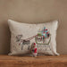 A square beige linen throw pillow with an embroidered Christmas scene of a reindeer pulling a sleigh filled with presents, displayed on a wooden surface against a textured beige wall.