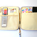An open canvas art bag organizer with a light beige interior, lavender zipper, and gold zipper pull. The left side has mesh pockets holding a watercolor palette, paintbrushes, and a color card, while the right side has a large pocket with watercolor swatches and a black case insert.