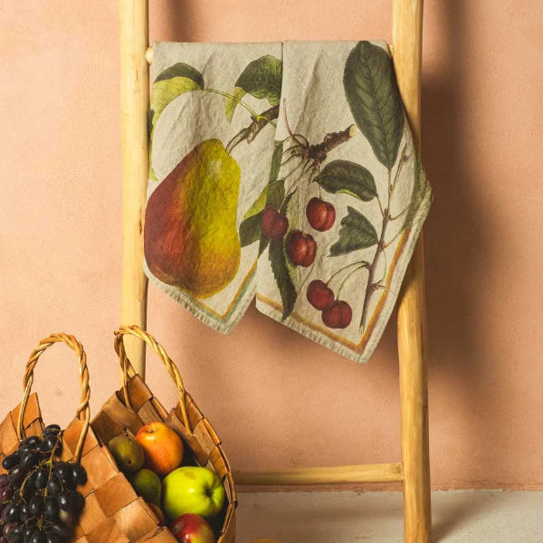 Decorative towel with fruit design on a wooden ladder against a pink wall, with a basket and fruits on the floor.