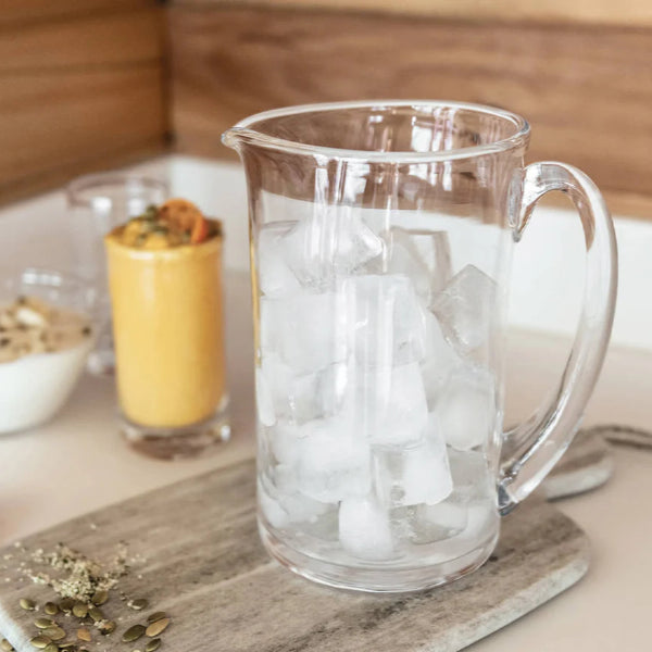 Clear glass pitcher filled with ice on a kitchen counter with a smoothie and oranges in the background.