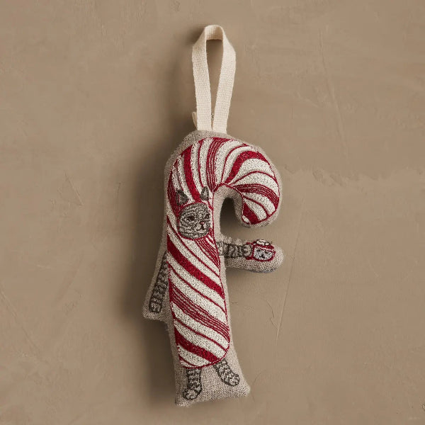 A festive, handw-embroidered Christmas ornament shaped like a candy cane, featuring red and white stripes with a gray cat walking along the side, hanging from a cream fabric loop against a textured beige background.