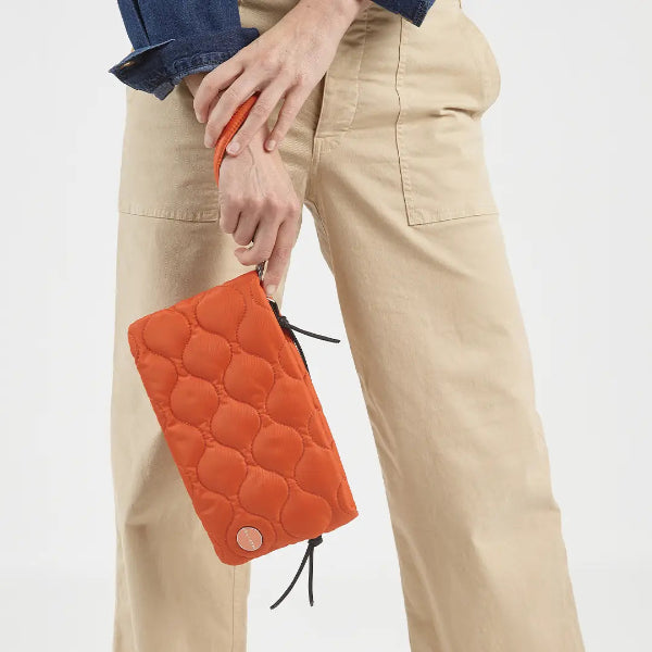A person wearing beige cargo pants and a blue denim shirt holds a bright orange quilted pouch with a matching orange handle against a clean white background.