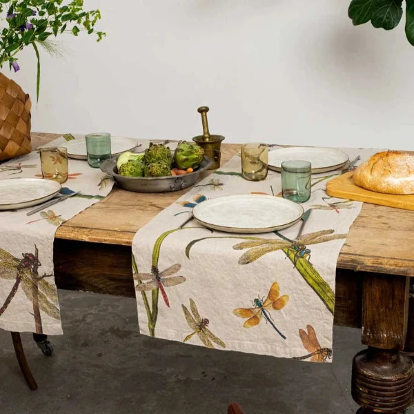 A linen table runner with a floral and dragonfly pattern is laid out on a wooden table, set with plates and cutlery for a meal.