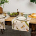 A linen table runner with a floral and dragonfly pattern is laid out on a wooden table, set with plates and cutlery for a meal.