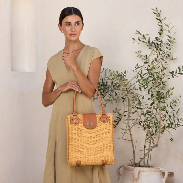 Woman holding a woven handbag in a neutral setting with a plant.