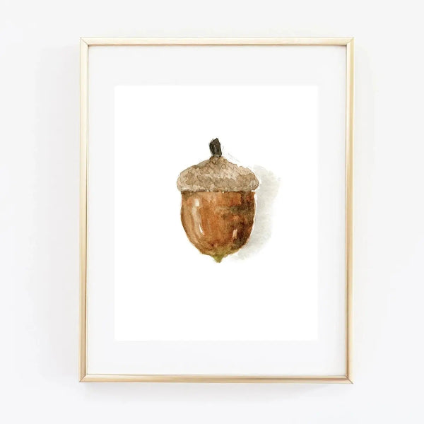Framed watercolor art print of a single brown acorn with a textured beige cap, set in a light wood frame with a white mat against a white wall.