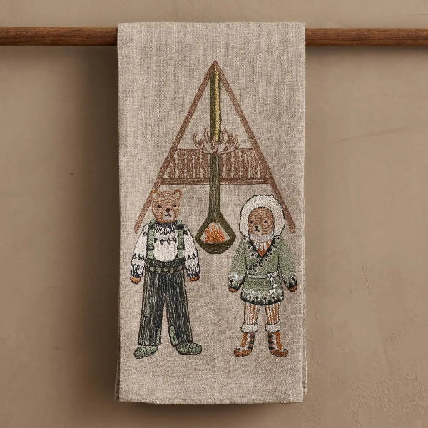 A beige linen tea towel hanging from a wooden rod against a warm beige wall. The towel features a detailed embroidered design of two children in winter clothing standing beneath a large triangular tent-like structure with a hanging spoon containing fire.