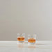 Two clear rocks glasses filled with an amber drink, placed on a light marble surface against a textured off-white wall.