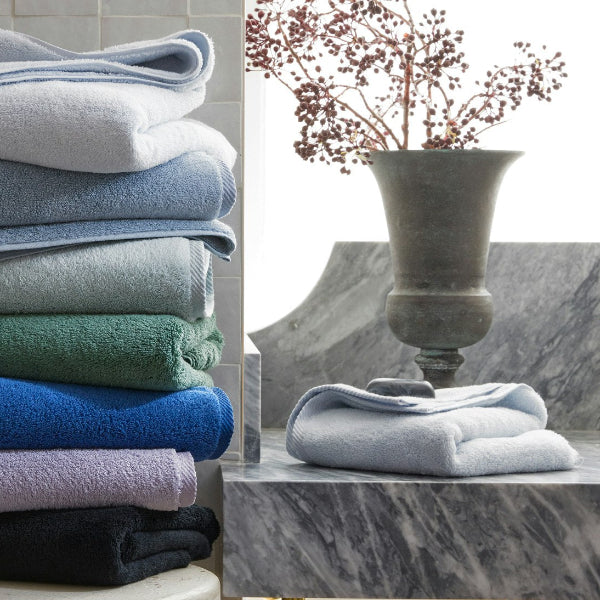 Stack of folded towels in various colors on a white surface with a vase in the background.