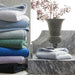 Stack of folded towels in various colors on a white surface with a vase in the background.