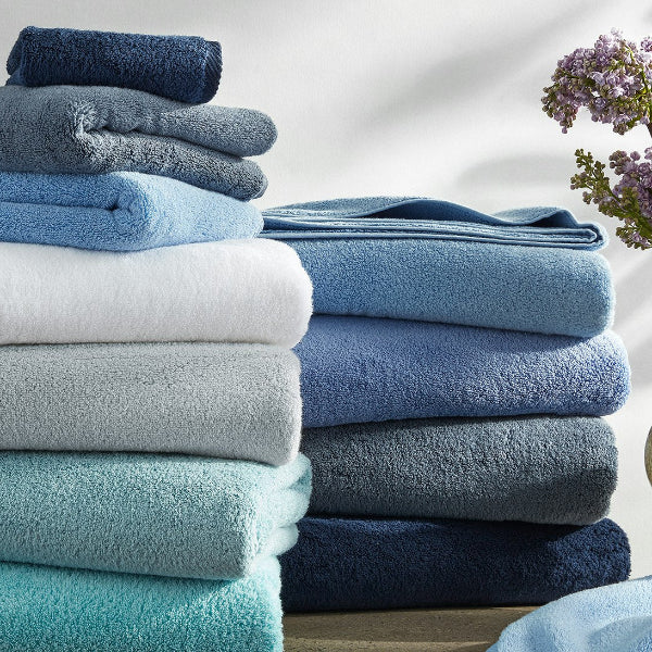 Stack of folded towels in various shades of blue and gray on a light background with a vase of flowers.