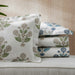 A stack of patterned bedding, including a large pillowcase and folded sheets, in white with green and blue botanical motifs, styled on a light beige sofa with a green plant leaf in the foreground.