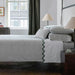 A neatly made bed in a styled bedroom, featuring a moss green and white patterned quilt, matching pillows, white sheets, and a dark green zigzag decorative trim. The room includes a dark wood canopy bed frame, bedside tables with lit lamps, a window with curtains, and a wooden floor.