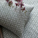 A close-up of a decorative throw pillow with a moss green trellis pattern on a white background, styled on a matching patterned bedspread with purple orchid flowers.