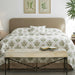 A neatly made bed in a bright bedroom setting, featuring a white duvet cover and matching pillow shams with a green botanical leaf pattern. The bed has a beige upholstered headboard, is flanked by dark wood nightstands with a lamp and books, and a matching bench sits at the foot of the bed.