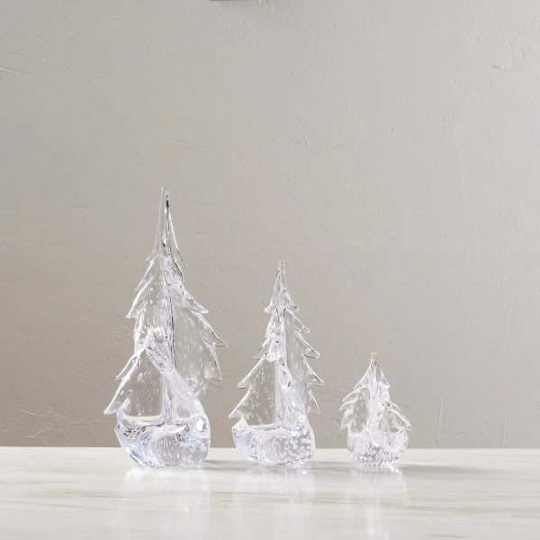 Three clear, textured Christmas tree ornaments of different sizes standing on a light marble surface against a neutral beige wall.