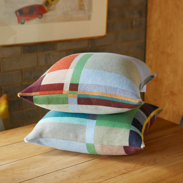 Two square throw pillows with a multicolor plaid pattern in muted shades of green, orange, blue, and burgundy, stacked on a wooden dining table in a cozy room setting.