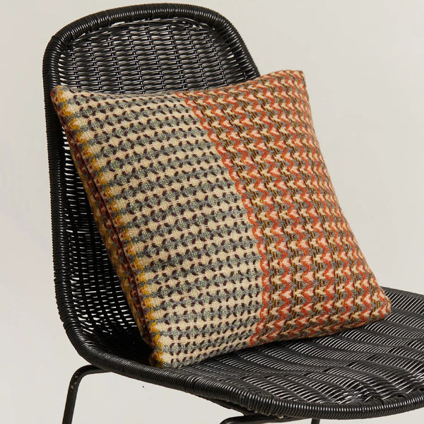 A square woven throw pillow with a geometric pattern in orange, beige, and black, resting on a black wicker chair against a plain light background.