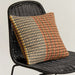 A square woven throw pillow with a geometric pattern in orange, beige, and black, resting on a black wicker chair against a plain light background.