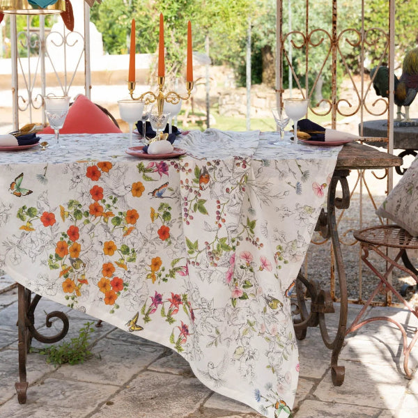 A floral tablecloth with orange, pink, and green botanical prints draped over a table under a white canopy, styled with metal garden chairs, orange taper candles, and colorful fabric garlands.