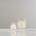 Two clear glass candle holders with footed bases, one small and one larger, each containing a lit cream candle, set on a light marble surface against a textured light gray wall.
