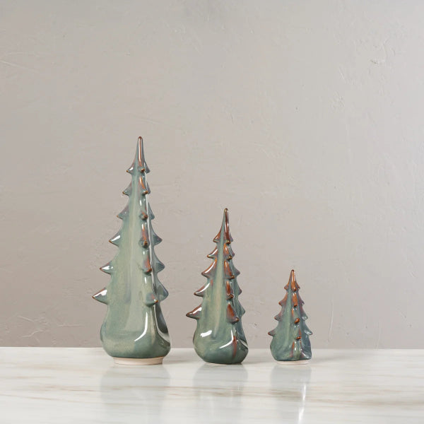 Three small ceramic Christmas tree ornaments in a glossy sage green glaze with metallic copper tips, displayed on a light marble surface against a neutral beige wall.