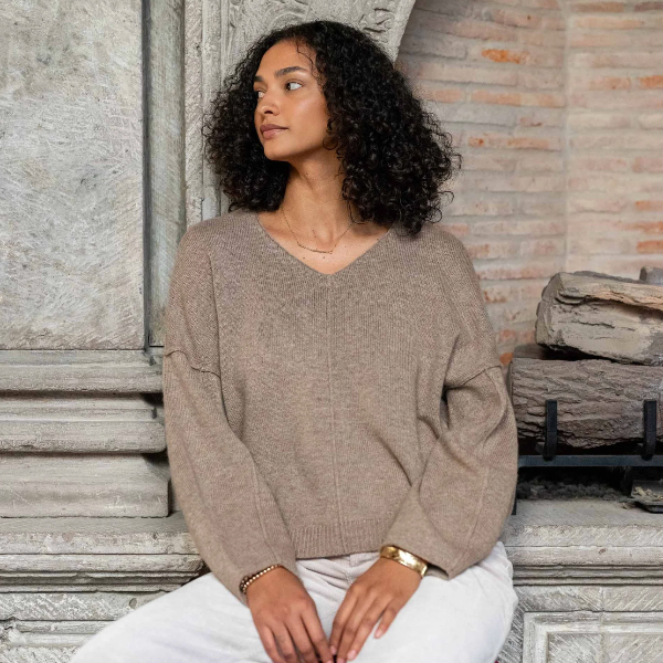 Woman wearing a beige sweater and white pants sitting against a stone wall.