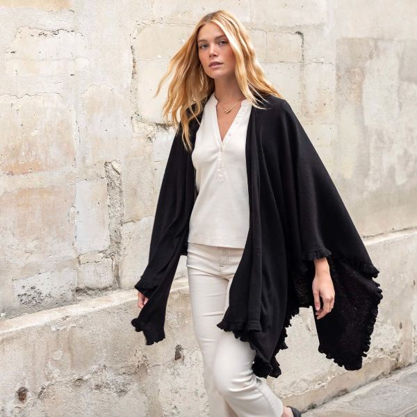 Woman wearing a black cardigan over a white top and pants against a stone wall.