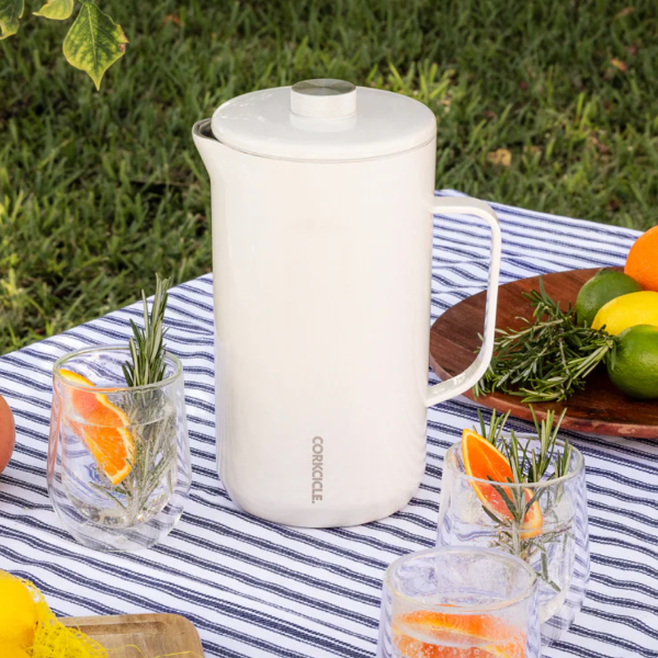 White pitcher on a striped tablecloth with glasses of orange slices and herbs outdoors.