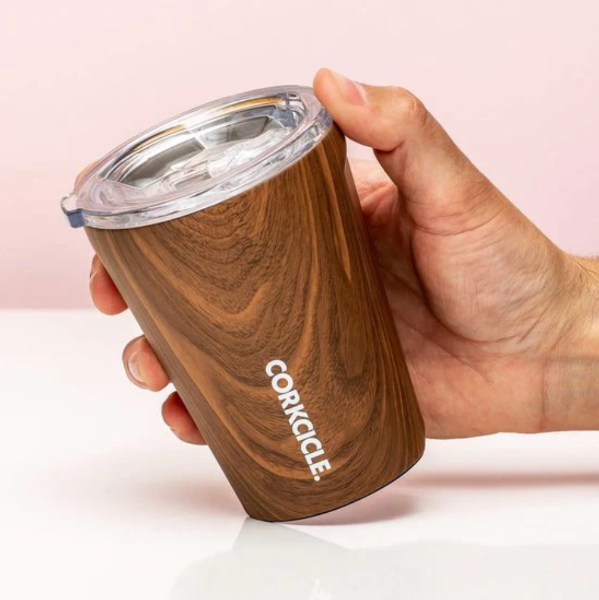 Wooden tumbler with clear lid held by a hand on a light pink background, featuring the Corkcicle brand.
