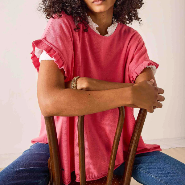 Person wearing a pink shirt with ruffled sleeves and blue jeans sitting on a wooden stool.