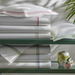 Stack of white bed sheets with decorative stripes on a green table with a plant in the background