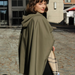 A woman wearing an olive green hooded cape-style coat stands on a cobblestone street, holding a beige plaid scarf. Modern buildings are visible in the background.