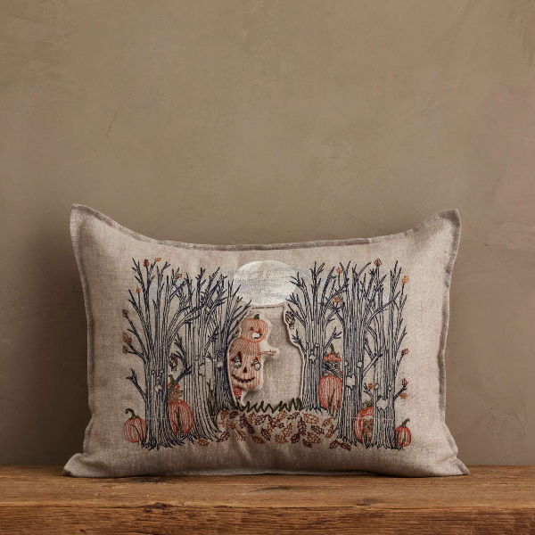 A square beige linen throw pillow with a rustic embroidered design of a pumpkin patch under a full moon, resting on a wooden surface against a textured beige wall.