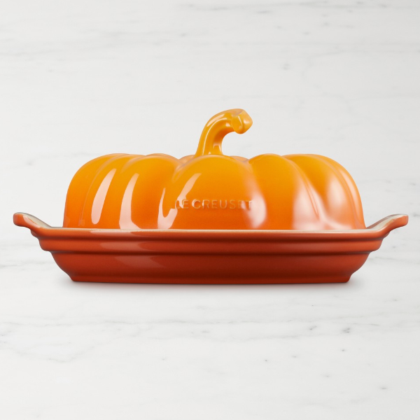 A Le Creuset pumpkin-shaped butter dish with a glossy orange lid and a reddish-orange rectangular base, photographed on a light marble surface.